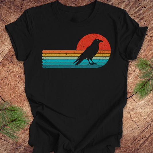 A black short-sleeve t-shirt laid flat on a rustic wood surface, featuring a distressed retro sunset graphic with horizontal stripes in orange, yellow, and blue, and a black raven silhouette standing in front of the sun.