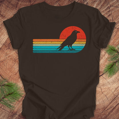 A dark brown t-shirt laid flat on a rustic wood background with pine branches in the corners. The shirt features a graphic of a black raven silhouette standing in front of a retro sunset with horizontal stripes in red, orange, yellow, and blue.