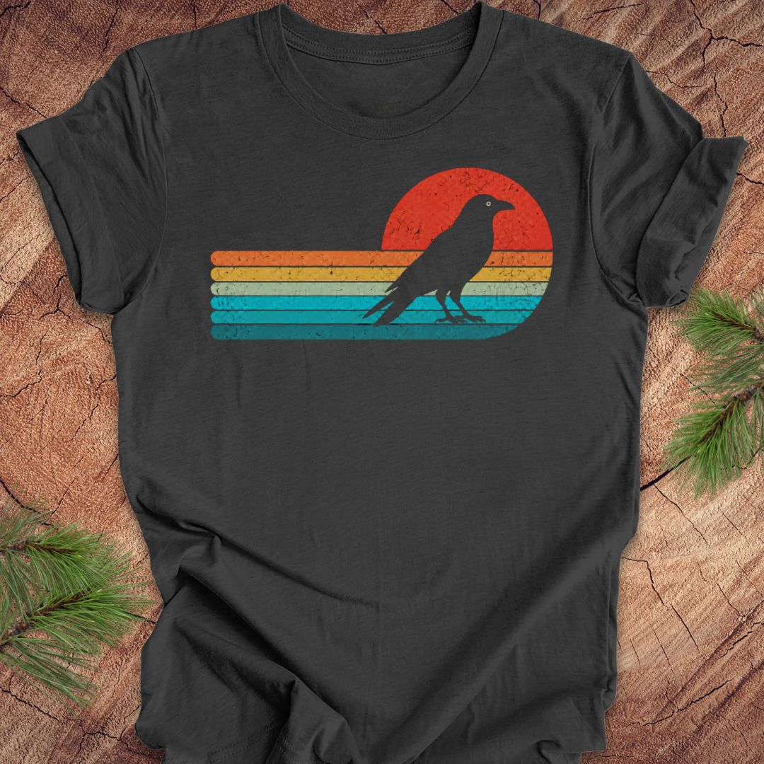 A dark charcoal gray t-shirt laid flat on a rustic wood background with pine branches in the corners. The shirt features a distressed retro graphic of a black bird silhouette standing in front of a sunset with horizontal stripes in red, orange, yellow, and blue.