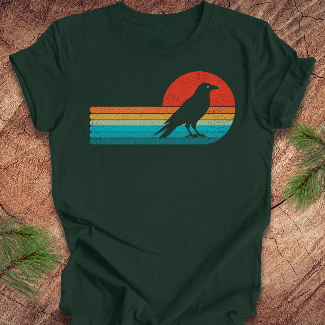 A dark forest green t-shirt featuring a retro sunset graphic with a black raven silhouette, laid flat on a rustic wood background with pine branches.