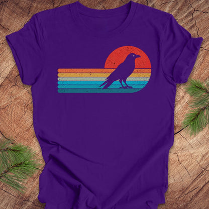 A purple short-sleeve crewneck t-shirt featuring a retro sunset graphic with a raven silhouette, laid flat on a rustic wood background with pine branches.
