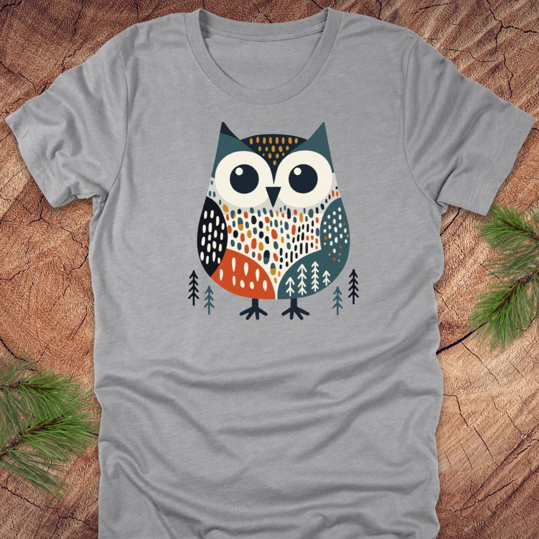 Gray t-shirt with a colorful owl design on a wooden surface with pine branches.