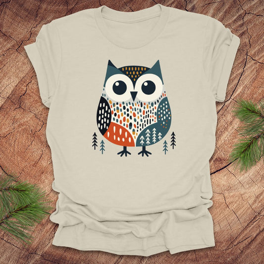 Beige t-shirt with a colorful owl design on a wooden surface with pine branches.