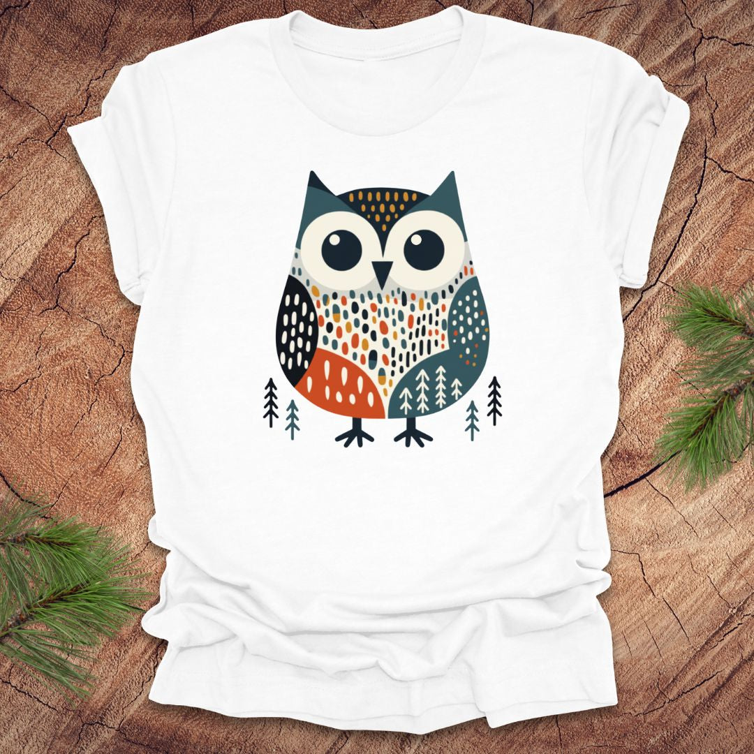 White t-shirt with an owl design on a wooden surface with pine branches.