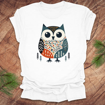 White t-shirt with an owl design on a wooden surface with pine branches.