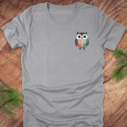 Gray t-shirt with a colorful pocket owl design on a wooden surface with pine branches.