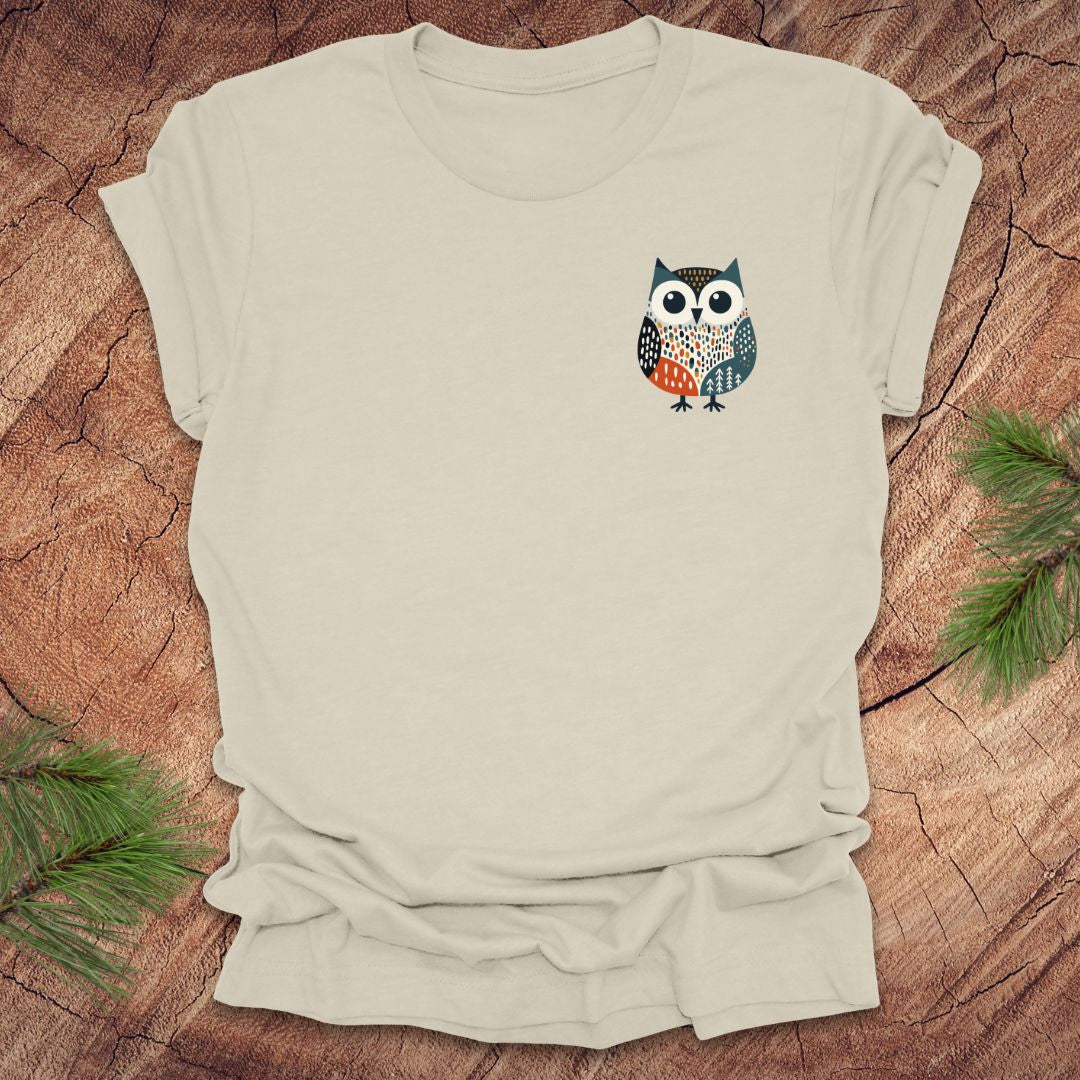 Beige t-shirt with a pocket owl design on a wooden surface with green leaves.