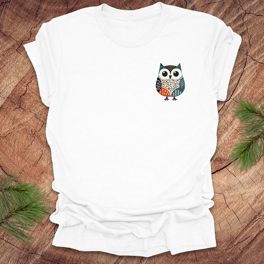 White t-shirt with a pocket owl design on a wooden surface with green leaves.