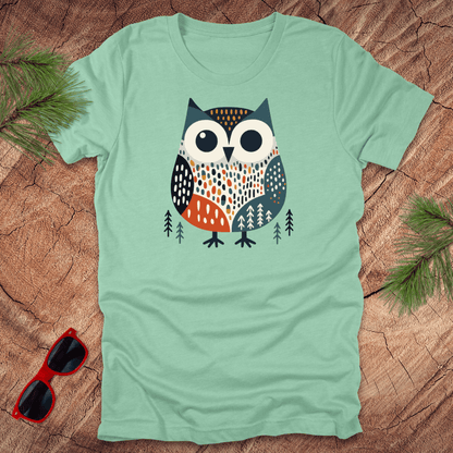 Mint green t-shirt with an owl design on a wooden surface with sunglasses and pine branches.