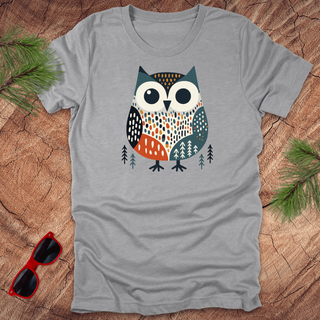 Gray t-shirt with a colorful owl design on a wooden surface with sunglasses and pine branches.