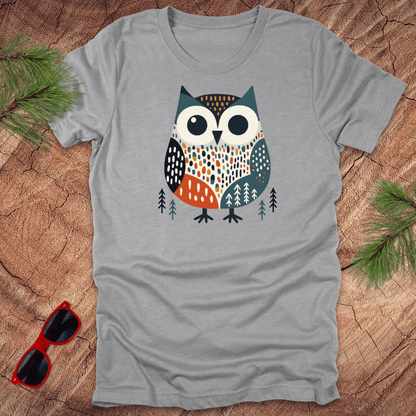 Gray t-shirt with a colorful owl design on a wooden surface with sunglasses and pine branches.
