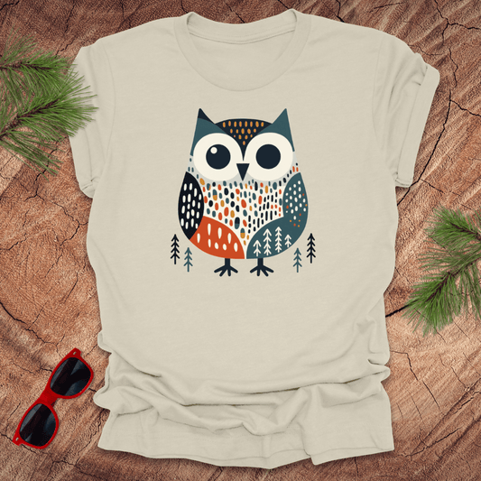 Beige t-shirt with an owl design on a wooden surface with sunglasses and pine branches.