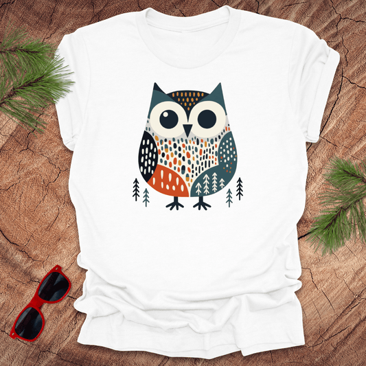 White t-shirt with a colorful owl design on a wooden surface with sunglasses and pine branches.