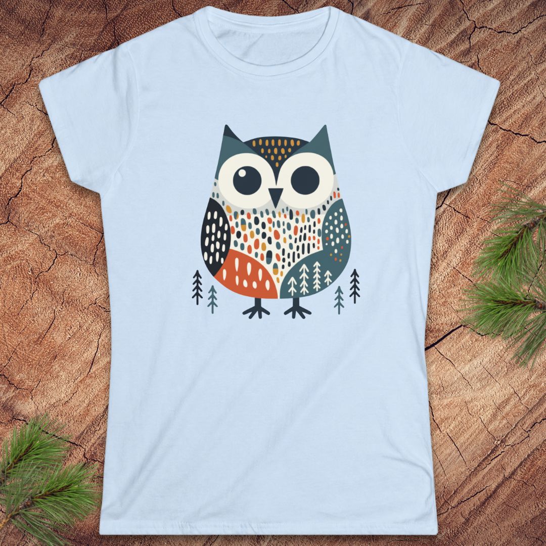 A light blue short-sleeve crewneck t-shirt featuring a stylized patterned owl graphic on the chest, laid flat on a rustic wood background with pine needles in the corners.