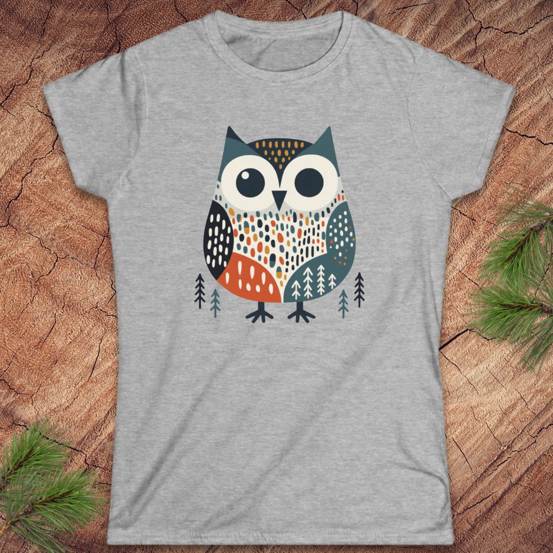 A heather gray short-sleeve t-shirt featuring a stylized owl graphic in teal, orange, black, and white, laid flat on a rustic wood surface with pine branches in the corners.