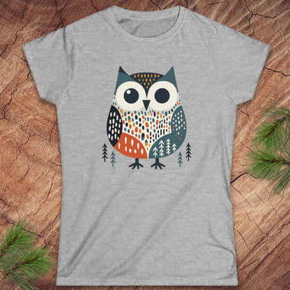 A heather gray short-sleeve t-shirt featuring a stylized owl graphic in teal, orange, black, and white, laid flat on a rustic wood surface with pine branches in the corners.