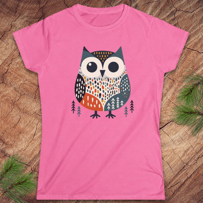 A pink short-sleeve crewneck t-shirt featuring a stylized graphic of an owl with geometric patterns in orange, white, black, and gray, surrounded by small pine trees, laid flat on a rustic wood background with pine needles.
