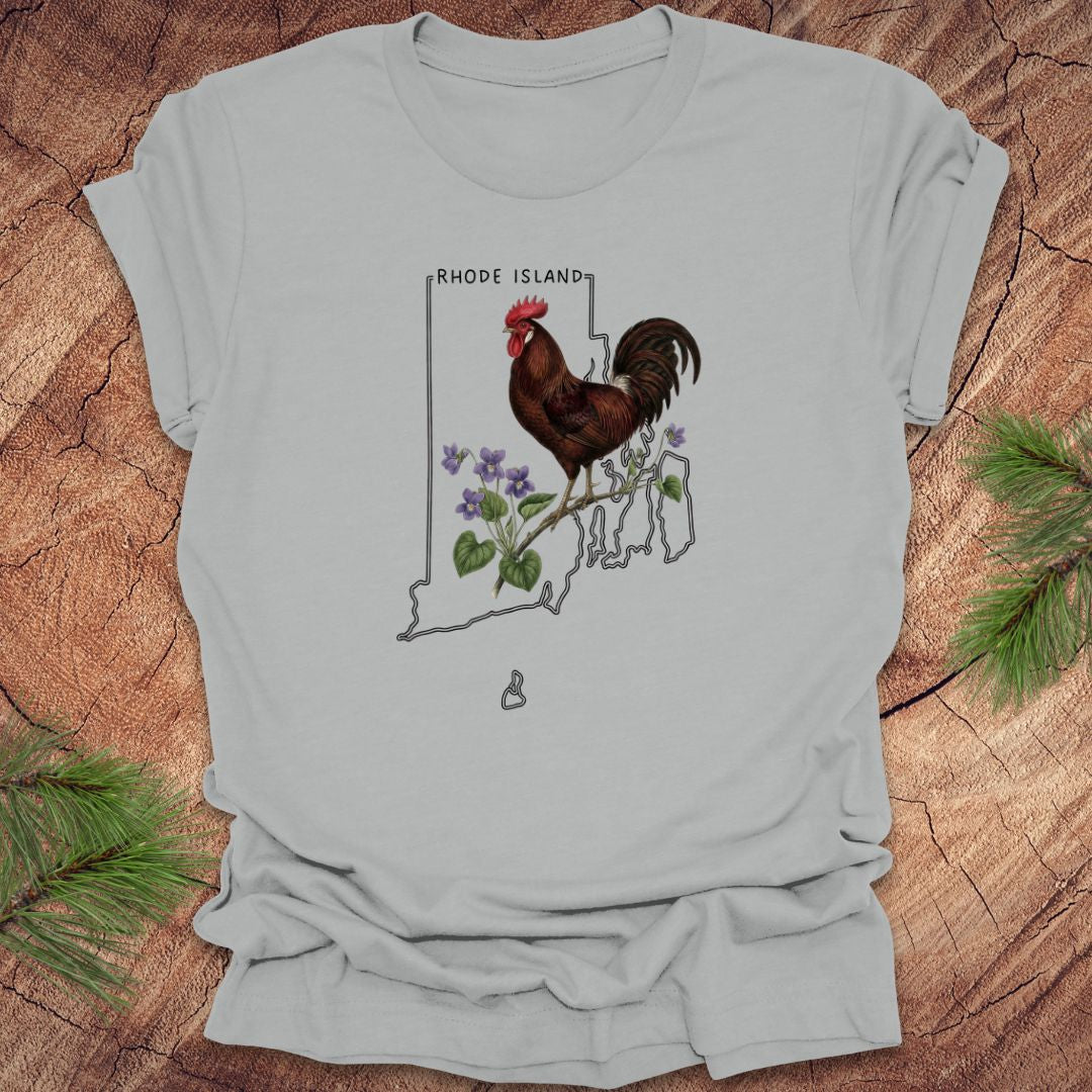 Silver t-shirt with the state outline of Rhode Island with state bird, red rooster and flower, violet,  on a wooden surface