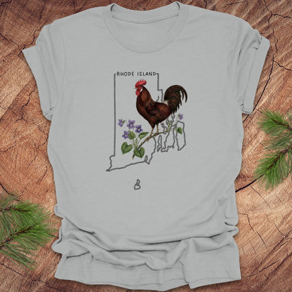 Silver t-shirt with the state outline of Rhode Island with state bird, red rooster and flower, violet,  on a wooden surface
