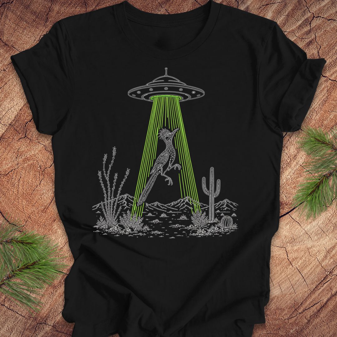 A black short-sleeve t-shirt laid flat on a rustic wood surface, featuring a graphic of a gray alien UFO casting a bright green beam of light that illuminates a gray kangaroo in a desert landscape with cacti.