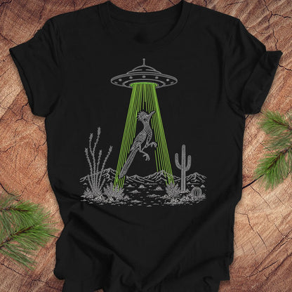 A black short-sleeve t-shirt laid flat on a rustic wood surface, featuring a graphic of a gray alien UFO casting a bright green beam of light that illuminates a gray kangaroo in a desert landscape with cacti.