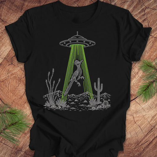 A black short-sleeve t-shirt laid flat on a rustic wood surface, featuring a graphic of a gray alien UFO casting a bright green beam of light that illuminates a gray kangaroo in a desert landscape with cacti.