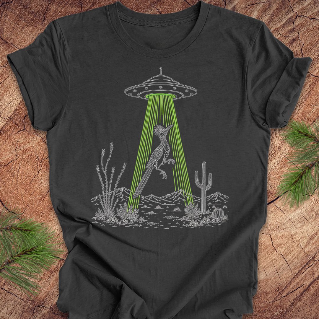 A dark charcoal gray t-shirt with a graphic of a gray alien spaceship emitting green laser beams at a desert fox, surrounded by cacti. The t-shirt is displayed flat on a rustic wood background with pine needles in the corners.