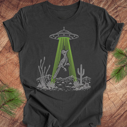 A dark charcoal gray t-shirt with a graphic of a gray alien spaceship emitting green laser beams at a desert fox, surrounded by cacti. The t-shirt is displayed flat on a rustic wood background with pine needles in the corners.