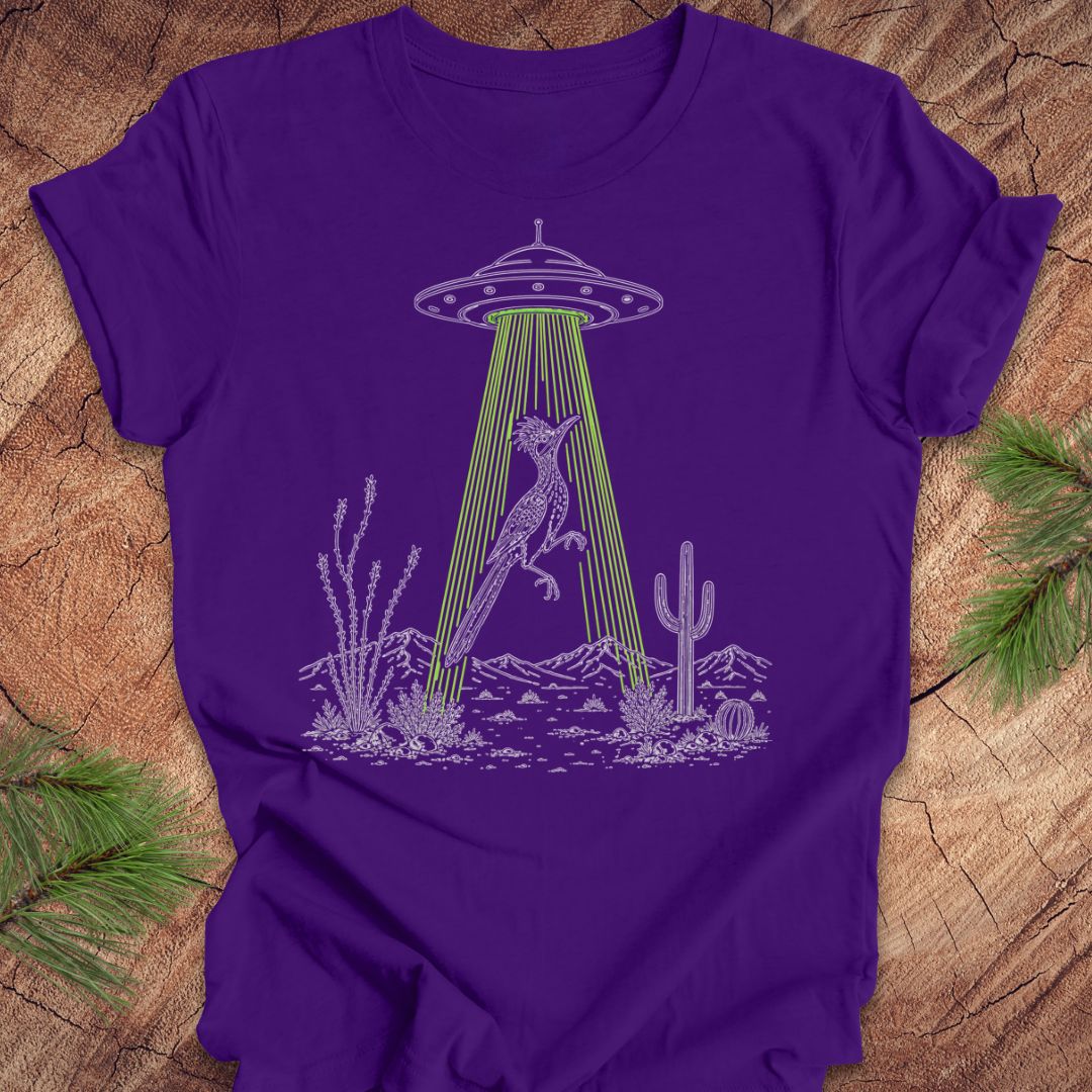 A purple short-sleeve t-shirt with a graphic of a green UFO beam striking a cat in a desert landscape, laid flat on a rustic wood background with pine branches.