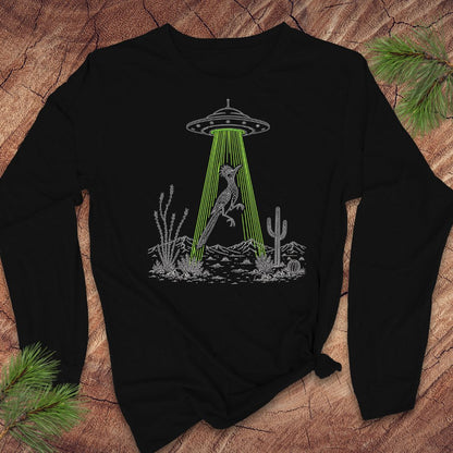 A black long-sleeve t-shirt with a graphic of a bird under a UFO in a desert scene, laid flat on a rustic wood background with pine branches.