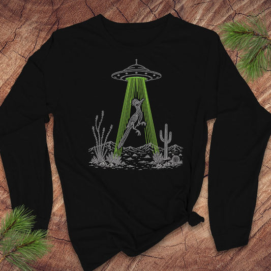 A black long-sleeve t-shirt with a graphic of a bird under a UFO in a desert scene, laid flat on a rustic wood background with pine branches.