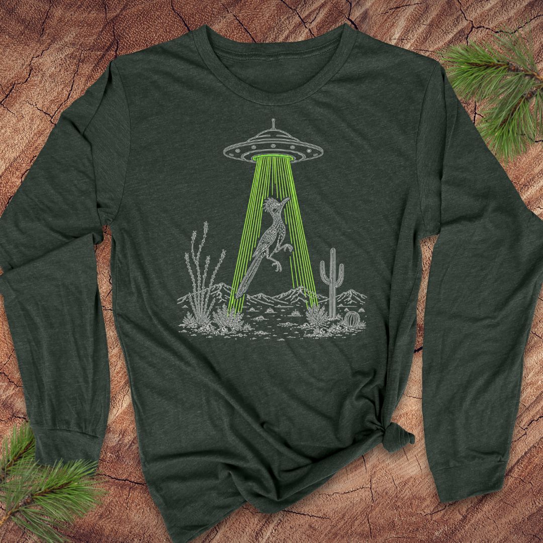 A green long-sleeve t-shirt with a graphic of a bird under a UFO in a desert scene, laid flat on a rustic wood background with pine branches.