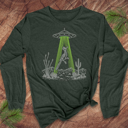 A green long-sleeve t-shirt with a graphic of a bird under a UFO in a desert scene, laid flat on a rustic wood background with pine branches.