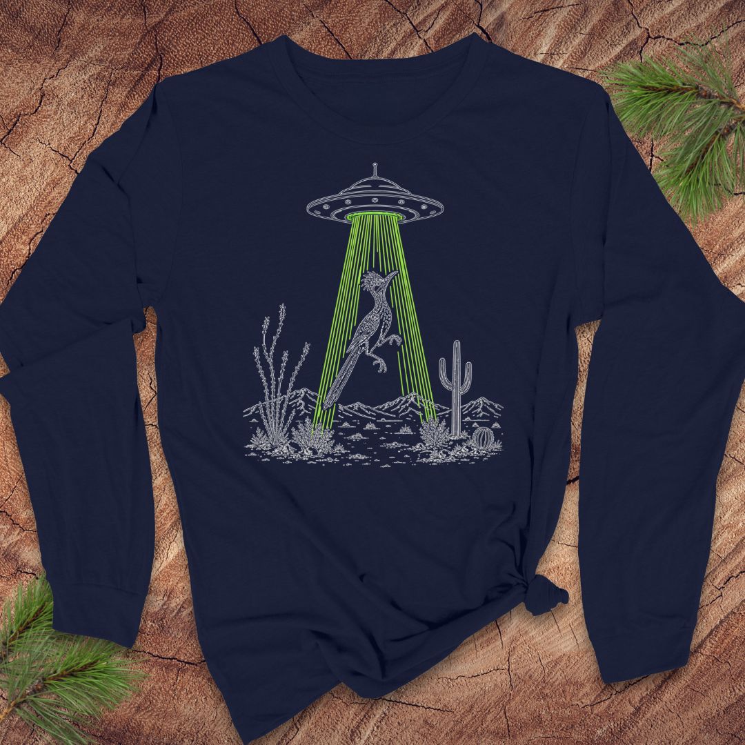 A navy blue long-sleeve t-shirt with a graphic of a bird under a UFO in a desert scene, laid flat on a rustic wood background with pine branches.