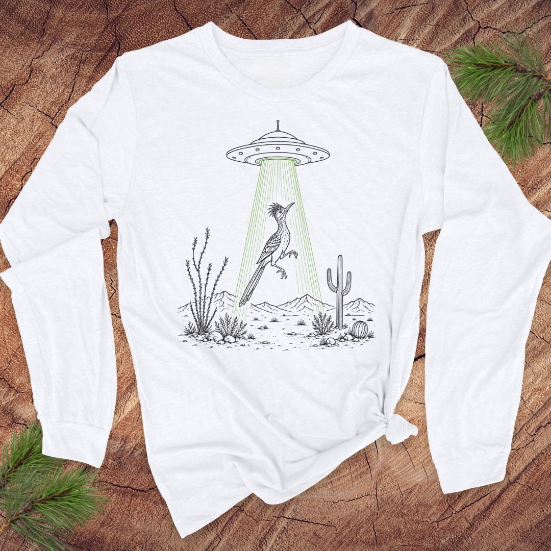 A white long-sleeve t-shirt with a graphic of a bird under a UFO in a desert scene, laid flat on a rustic wood background with pine branches.