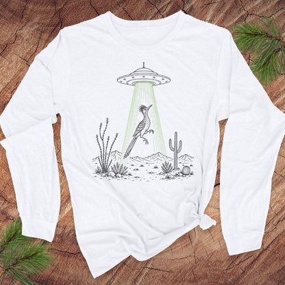 A white long-sleeve t-shirt with a graphic of a bird under a UFO in a desert scene, laid flat on a rustic wood background with pine branches.