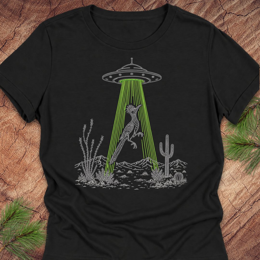 A black short-sleeve crewneck t-shirt laid flat on a rustic wood background with pine branches in the corners. The shirt features a graphic print of a gray desert scene with a cactus, a rabbit, and a UFO emitting bright green laser beams.