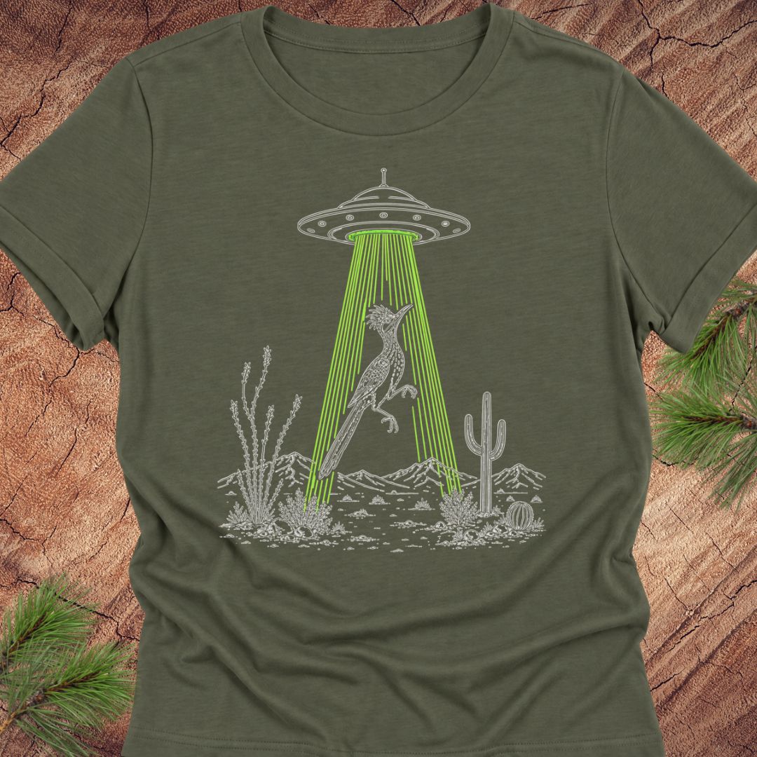 An olive green short-sleeve t-shirt with a graphic print of a gray desert scene featuring a cactus, a rabbit under a UFO with bright green beams, and pine branches in the corners, set against a wood-grain background.