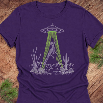A purple short-sleeve crewneck t-shirt laid flat on a rustic wood background with pine branches in the corners. The shirt features a graphic print of a desert coyote under a UFO with green laser beams.