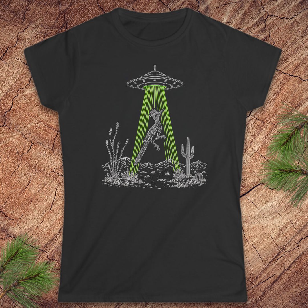 A black short-sleeve crewneck t-shirt laid flat on a textured wood background with pine branches in the corners. The shirt features a black line art graphic of a bird under a UFO with green rays shining down toward a desert landscape.