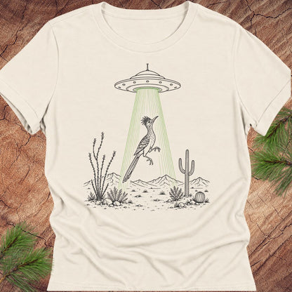An off-white short-sleeve crewneck t-shirt featuring a graphic print of a desert bird under a UFO with green beam rays, laid flat on a rustic wood background with pine branches.