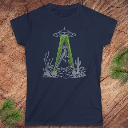 A navy blue short-sleeve crewneck t-shirt laid flat on a textured wood background with pine branches in the corners. The shirt features a black line art graphic of a bird under a UFO with green rays shining down toward a desert landscape.