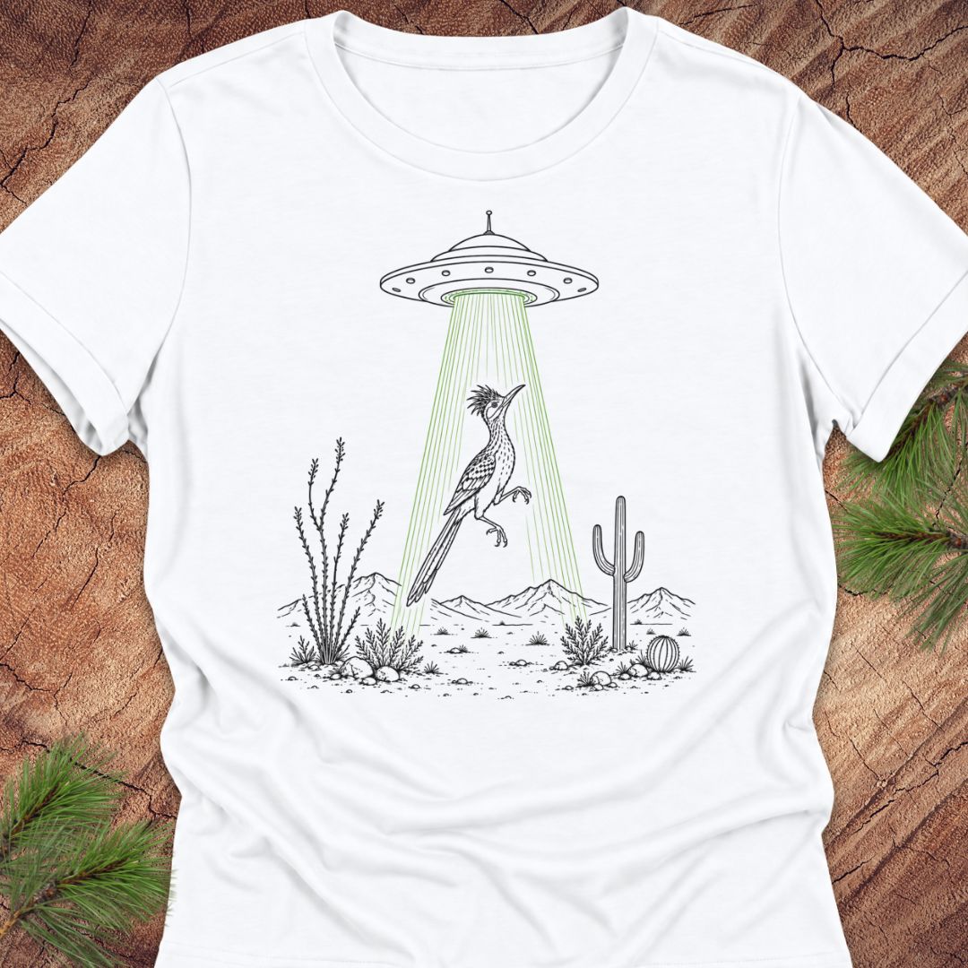 A white short-sleeve crewneck t-shirt laid flat on a rustic wood background with pine branches. The shirt features a graphic print of a black-and-white desert bird under a UFO with green light rays shining down.