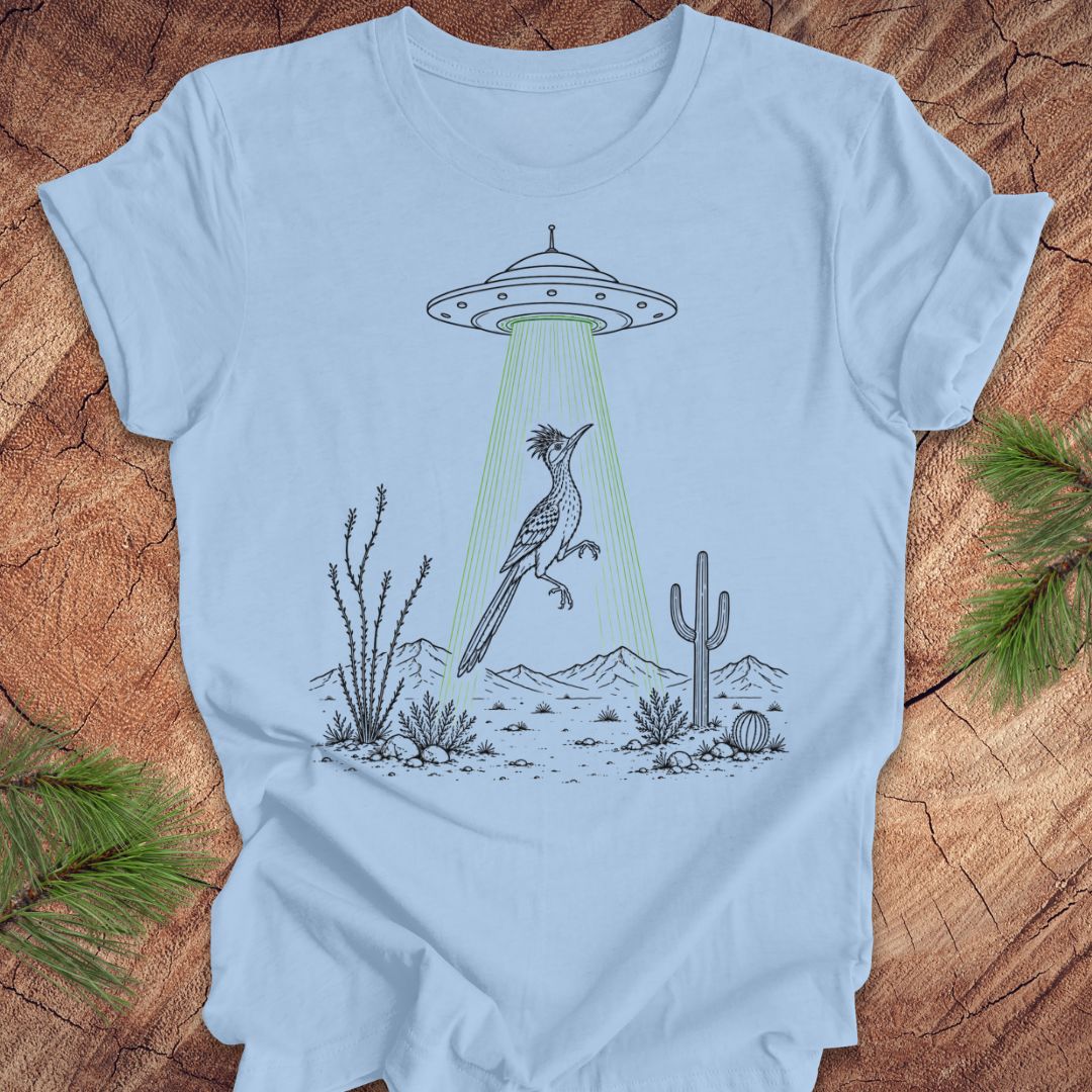 A light blue crewneck t-shirt laid flat on a rustic wood background with pine branches in the corners. The shirt features a graphic illustration of a desert scene with a UFO casting a green beam of light that hits a large crested bird.