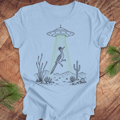 A light blue crewneck t-shirt laid flat on a rustic wood background with pine branches in the corners. The shirt features a graphic illustration of a desert scene with a UFO casting a green beam of light that hits a large crested bird.
