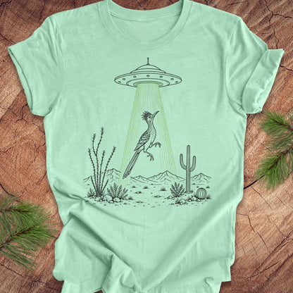A mint green graphic t-shirt laid flat on a rustic wood background with pine branches in the corners. The t-shirt features a black line art illustration of a bird under a UFO with green rays shining down in a desert scene.