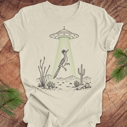 An off-white graphic t-shirt laid flat on a rustic wood background with pine branches. The t-shirt features a black line art illustration of a desert bird under a UFO with green rays shining down.