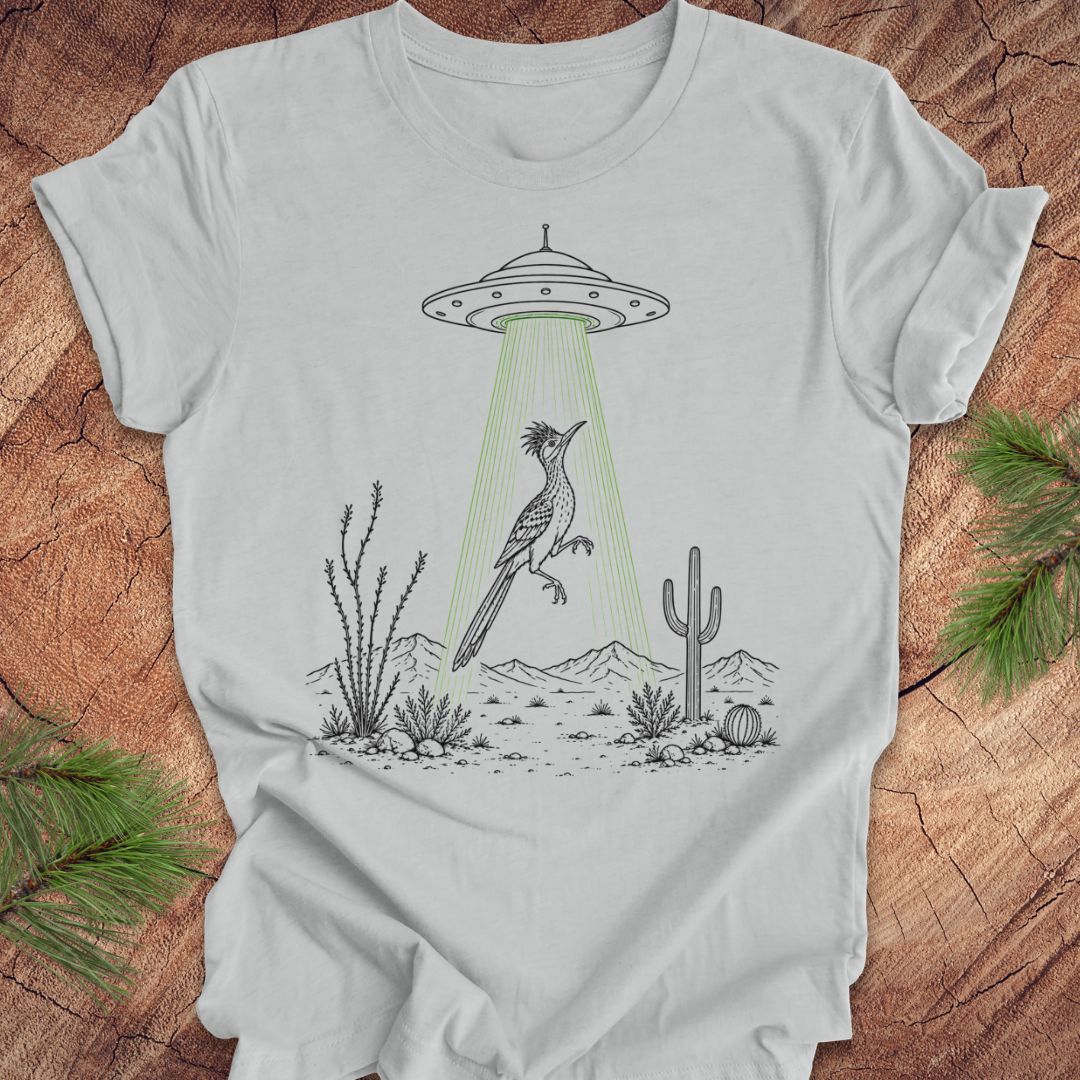 A light gray short-sleeve crewneck t-shirt with a graphic print of a gray bird under a green beam from a UFO, styled on a rustic wood background with pine branches.