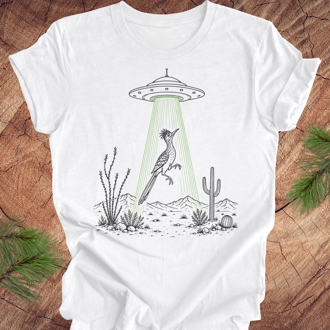 A white short-sleeve crewneck t-shirt laid flat on a rustic wood background with pine branches in the corners. The t-shirt features a black line art graphic of a desert scene with a flying bird under a green beam from a UFO.