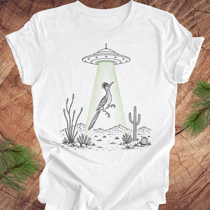 A white short-sleeve crewneck t-shirt laid flat on a rustic wood background with pine branches in the corners. The t-shirt features a black line art graphic of a desert scene with a flying bird under a green beam from a UFO.